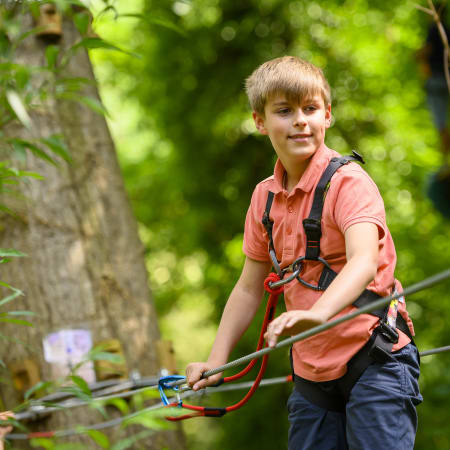 Adolescent en parcours aventure équipé de harnais et baudrier, accrochant sa corde à une poulie dans un camp sportif en forêt