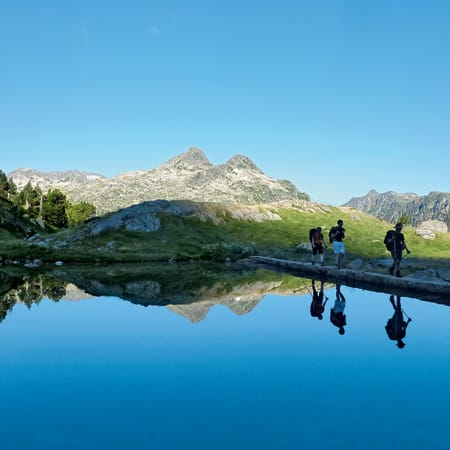 Randonneurs traversant un lac de montagne avec reflet des pics rocheux et forêts de conifères aux lacs de Néouvielle