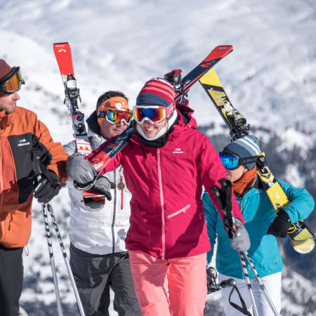 Groupe de quatre skieurs et snowboarders souriants à Serre Chevalier avec skis et snowboards levés, paysage montagneux enneigé en arrière-plan