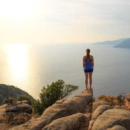 Jeune fille en tenue de sport sur rochers face à la mer de Corse au lever du soleil, montagnes en arrière-plan