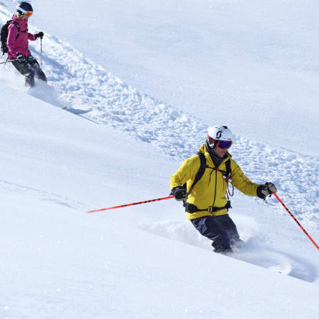 Skieur expert en veste jaune descendant une pente hors-piste enneigée avec moniteur en arrière-plan dans le Briançonnais