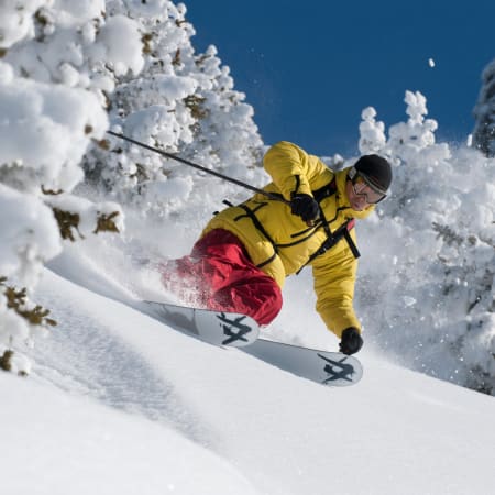 Skieur expert descendant une pente hors-piste en poudreuse fraîche entre sapins enneigés, vêtu d'une veste jaune et d'un pantalon rouge