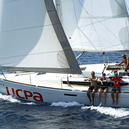 Voilier avec équipage naviguant en Méditerranée, voiles blanches déployées, passagers à bord en mer turquoise les pieds dans l'eau
