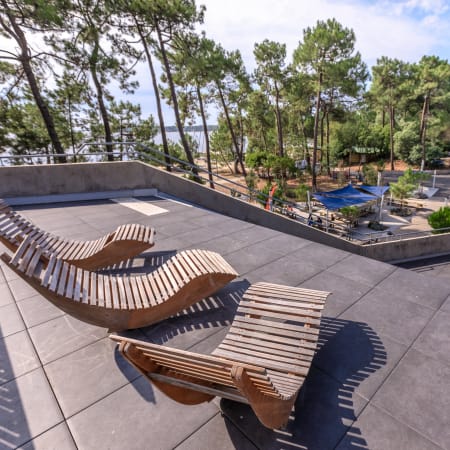 Terrasse avec chaises longues en bois face aux terrains de sport et lac de Bombannes entouré de pins