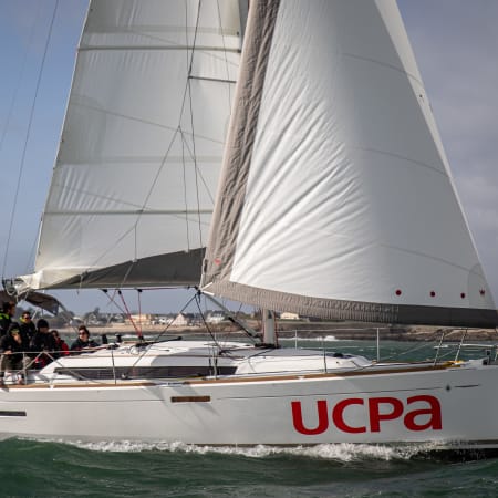 Voilier blanc UCPA naviguant en Morbihan avec équipage à bord et voiles gonflées, archipel côtier en arrière-plan