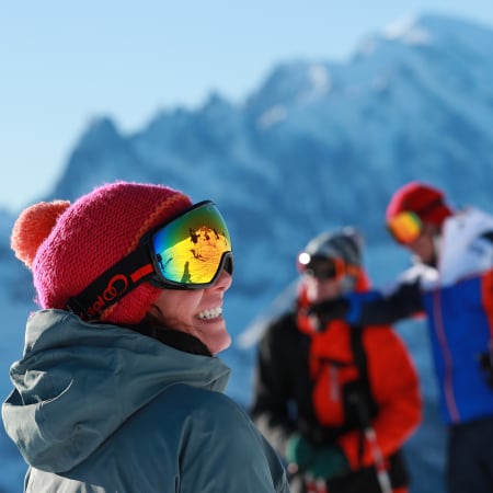 Jeune skieur en bonnet rose et masque de ski souriant face aux Deux-Alpes avec groupe d'amis en arrière-plan