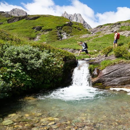 Randonneurs traversant un ruisseau alpin dans les prairies vertes de Serre Chevalier avec pics rocheux en arrière-plan