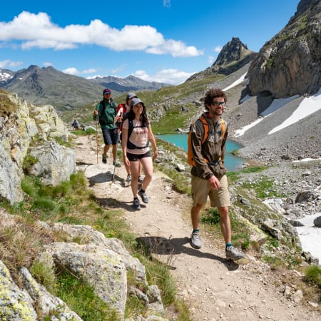 Randonneurs sur sentier d'altitude à Serre Chevalier avec lac turquoise, névés et chaîne de montagnes en arrière-plan