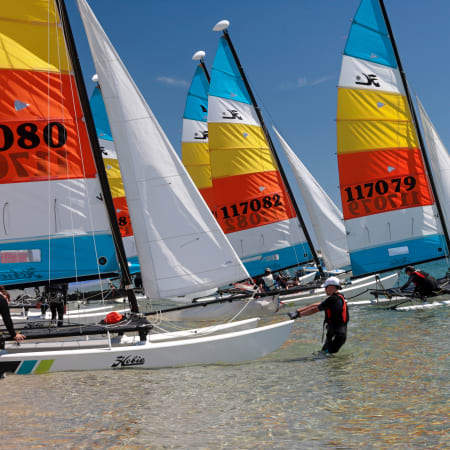 Catamarans avec voiles colorées alignés sur la plage, moniteur en combinaison préparant les embarcations pour la navigation en mer