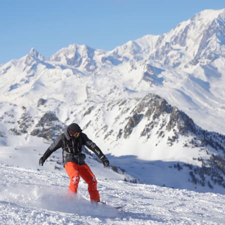Skieur descendant une piste enneigée avec vue sur des sommets alpins enneigés et ciel bleu dégagé
