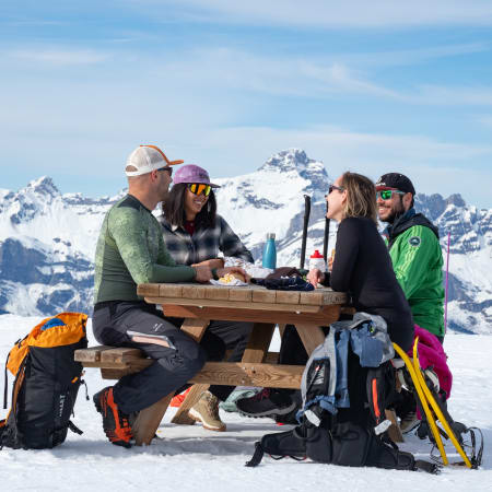 Groupe de randonneurs prenant une pause autour d'une table en montagne enneigée avec vue panoramique sur les Alpes