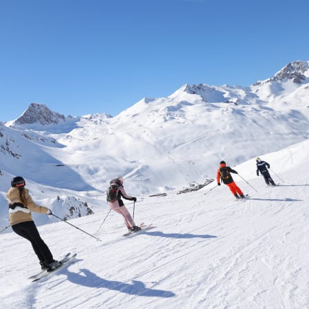 Skieurs progressant sur piste bleue enneigée en montagne avec sommets alpins en arrière-plan