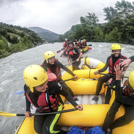 Groupe de jeunes en rafting sur le lac de Serre-Ponçon avec casques jaunes et gilets de sauvetage rouges, entouré de montagnes verdoyantes