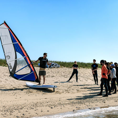 Cours de windsurf en groupe sur la plage avec voile bleue et rouge, moniteur et apprentis en combinaison