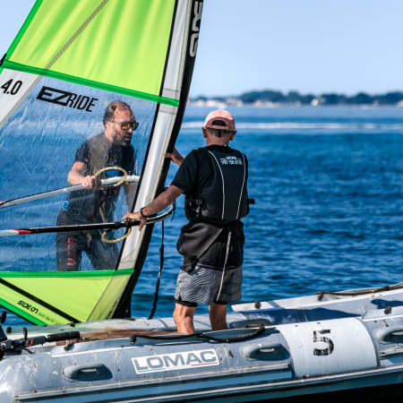 Moniteur de windsurf accompagnant un élève sur une planche avec voile verte en Bretagne face à l'océan