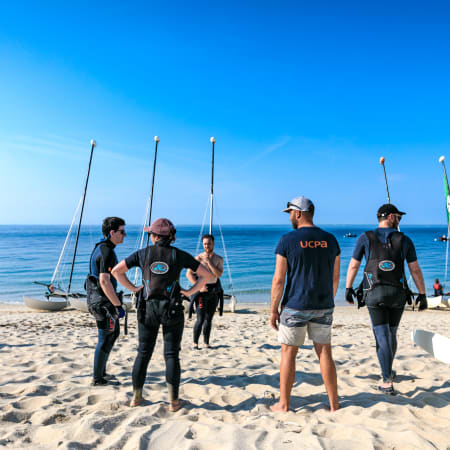 Groupe de jeunes en combinaison et équipement de catamaran sur la plage avec voiliers et archipel en arrière-plan