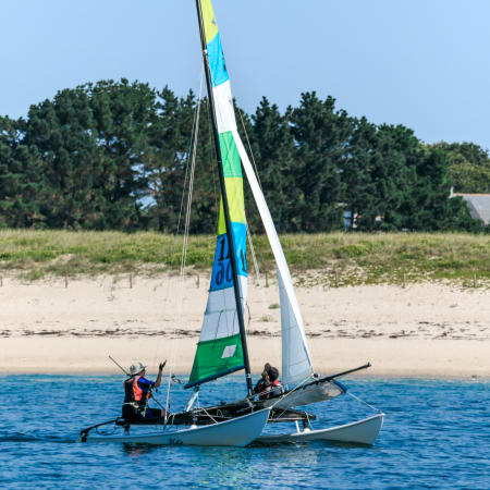 Catamaran à voile avec deux participants en mer, voile multicolore jaune et verte, plage et forêt en arrière-plan