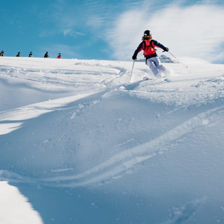 Skieur en veste rouge dévale une pente de poudreuse avec groupe de skieurs en arrière-plan dans un paysage montagneux enneigé