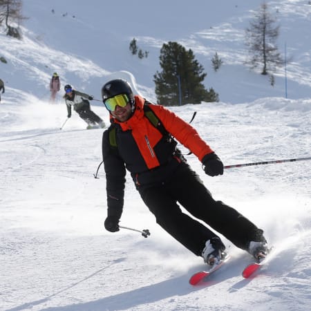 Skieur en veste rouge dévale une piste enneigée des Orres avec d'autres skieurs en arrière-plan et sapins alpins