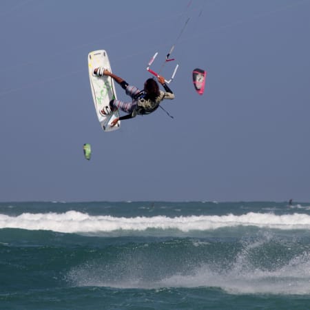 Kitesurfeur en vol effectuant un trick au-dessus des vagues avec plusieurs cerfs-volants visibles sur la plage de kitebeach