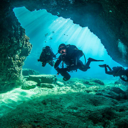 Plongeurs équipés de bouteilles explorant une grotte sous-marine avec rayons de lumière bleue traversant l'eau cristalline