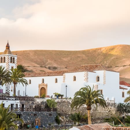 Église blanche aux volets dorés entourée de palmiers avec paysage volcanique aride en arrière-plan