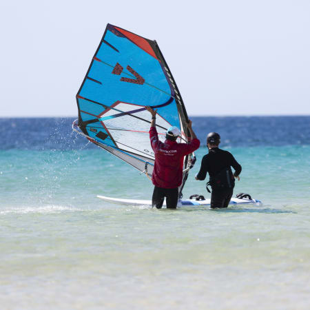 Deux pratiquants de windsurf en mer avec voile bleue et rouge, préparant leur sortie en eau turquoise et peu profonde