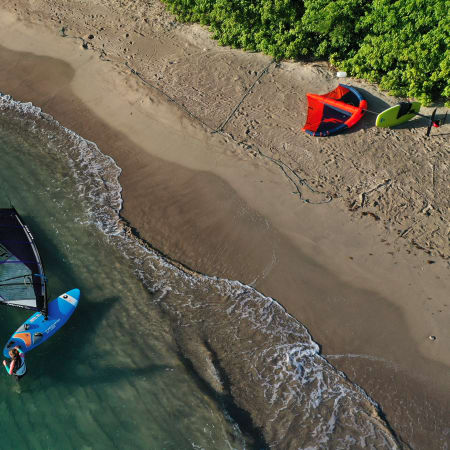Planche à voile et équipement de windsurf sur plage de sable avec eaux turquoises et végétation tropicale