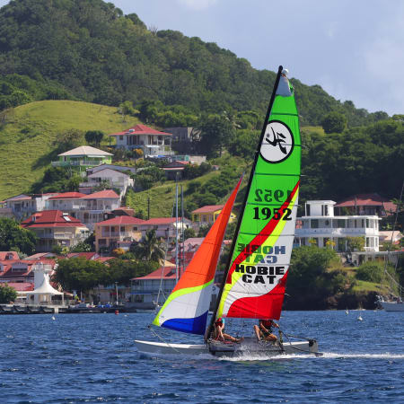 Catamaran aux voiles colorées naviguant dans la baie des Saintes avec îles verdoyantes et village côtier en arrière-plan