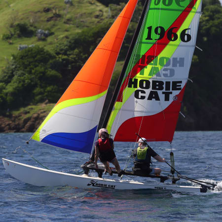 Catamaran avec voiles colorées naviguant en baie des Saintes, deux navigateurs à bord, paysage tropical verdoyant en arrière-plan