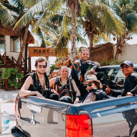 Groupe de plongeurs en tenue de plongée dans un véhicule à Playa del Carmen, palmiers et bâtiments coloniaux en arrière-plan