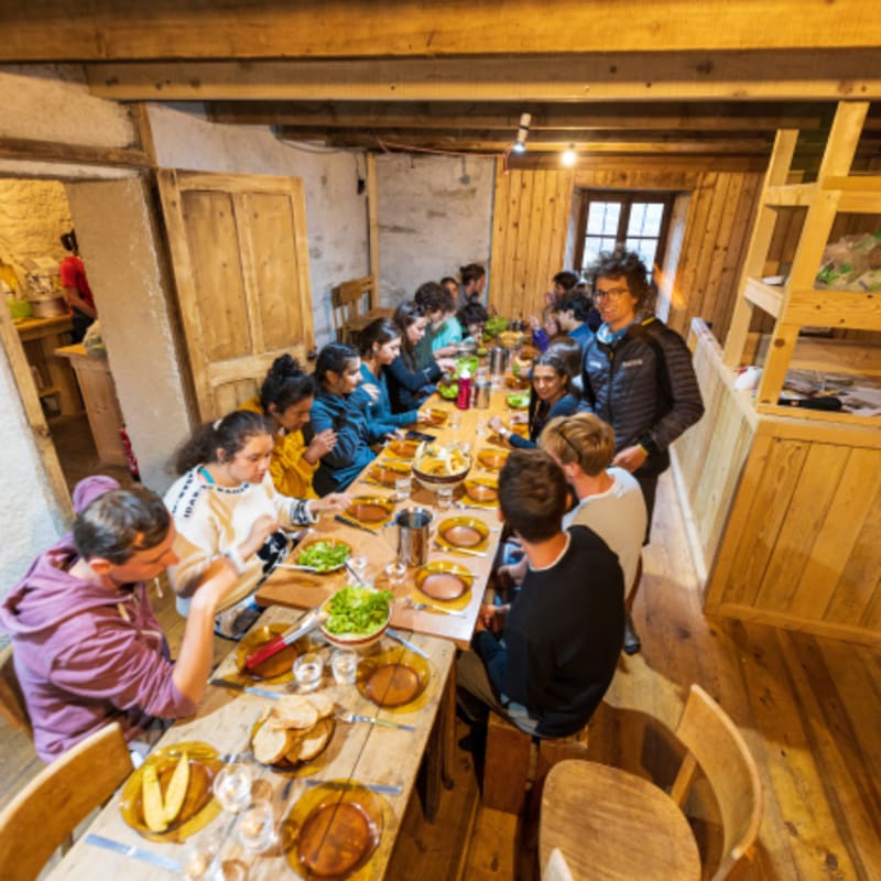 Jeunes randonneurs partageant un repas en refuge montagnard avec table en bois et lits superposés