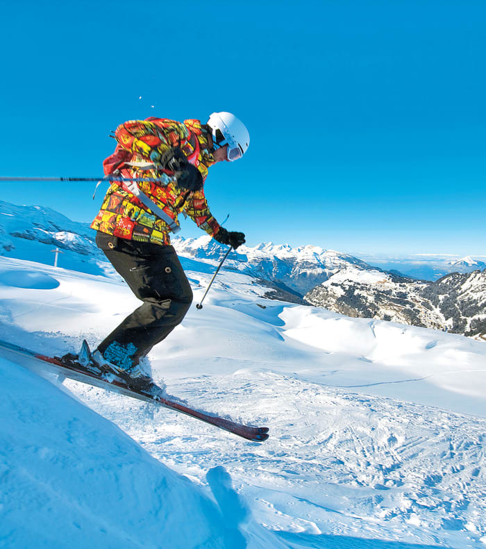Skieur en descente hors-piste sur pente enneigée avec montagnes alpines et ciel bleu dégagé