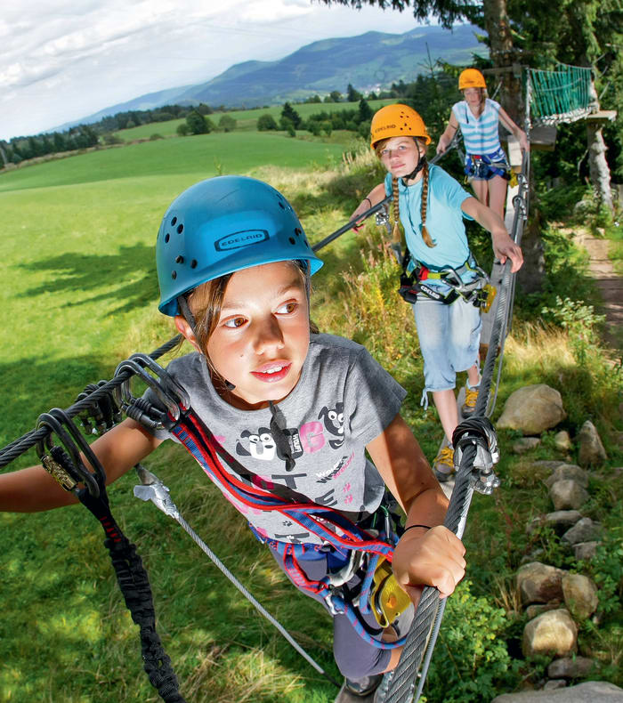 Enfants en via ferrata équipés de casques et harnais dans les Pyrénées avec vue sur vallée verdoyante