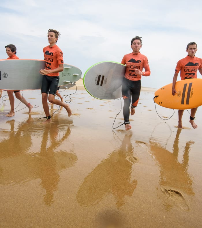 Groupe de jeunes surfeurs en maillots orange courant sur la plage bretonne avec leurs planches de surf