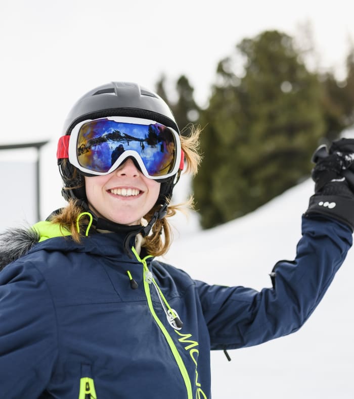 Jeune skieur débutant souriante aux Arcs portant casque et lunettes de ski, tenant ses bâtons sur piste enneigée
