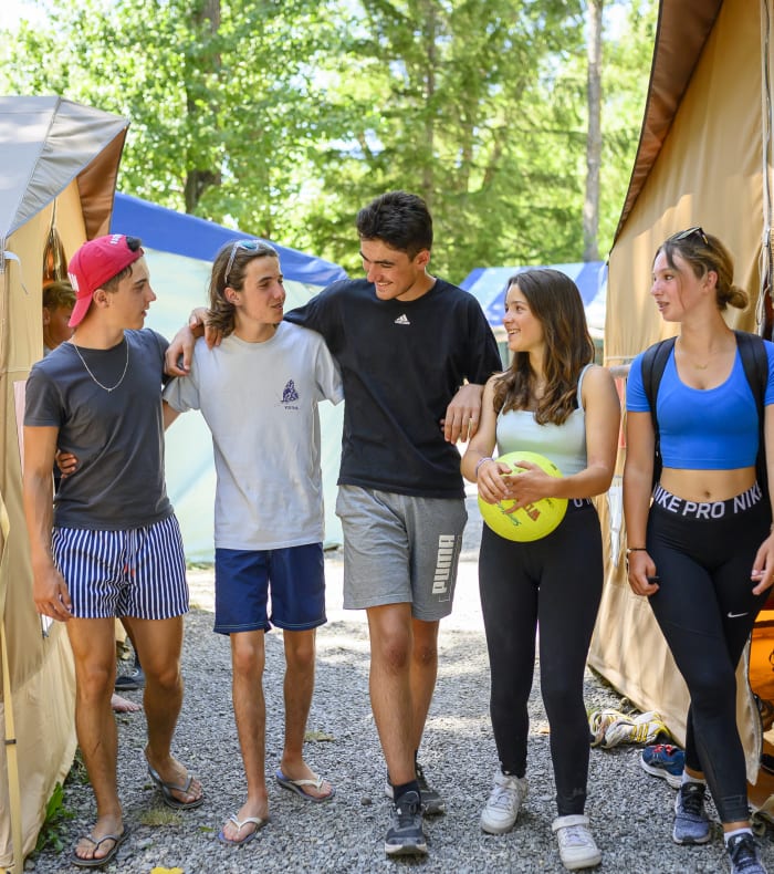 Groupe d'adolescents en tenue de sport discutant dans un campement avec tentes beiges en montagne