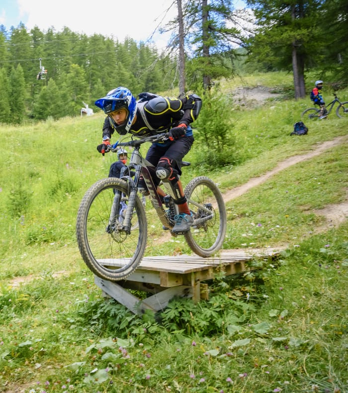 Cycliste en VTT sautant un module en bois dans un bikepark alpin avec forêt de sapins en arrière-plan