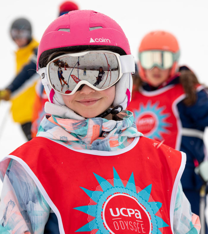 Enfant portant casque de ski rose et masque de ski souriant lors d'un séjour ski aux Orres avec d'autres jeunes skieurs en arrière-plan