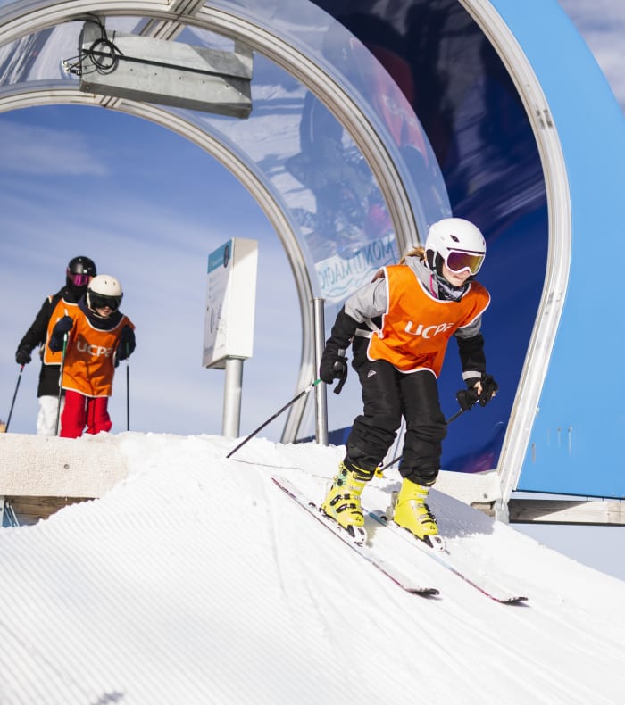 Enfant en tenue de ski orange descendant une piste avec remontée mécanique et autres skieurs en arrière-plan