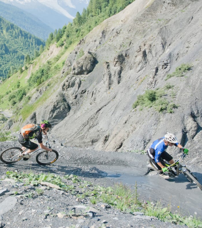 Trois vététistes roulant sur un sentier de scree aux pentes rocheuses du Mont-Blanc avec forêt alpine en arrière-plan