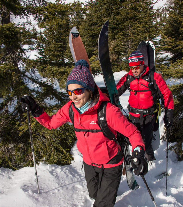Deux randonneurs en splitboard portant vestes rouges dans la vallée enneigée avec forêt de mélèzes en arrière-plan