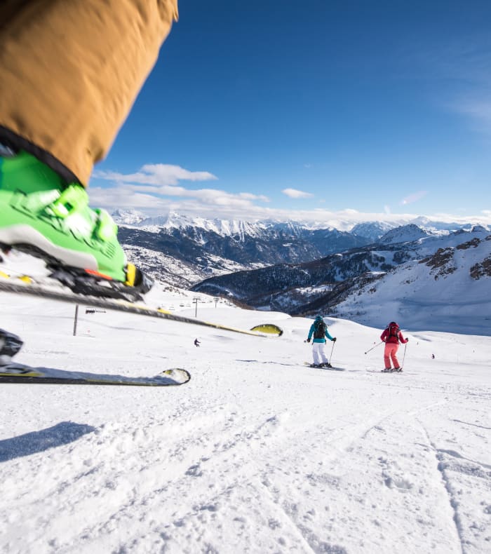 Skieurs en ski alpin dévallant une piste enneigée avec montagnes alpines en arrière-plan et remontées mécaniques visibles