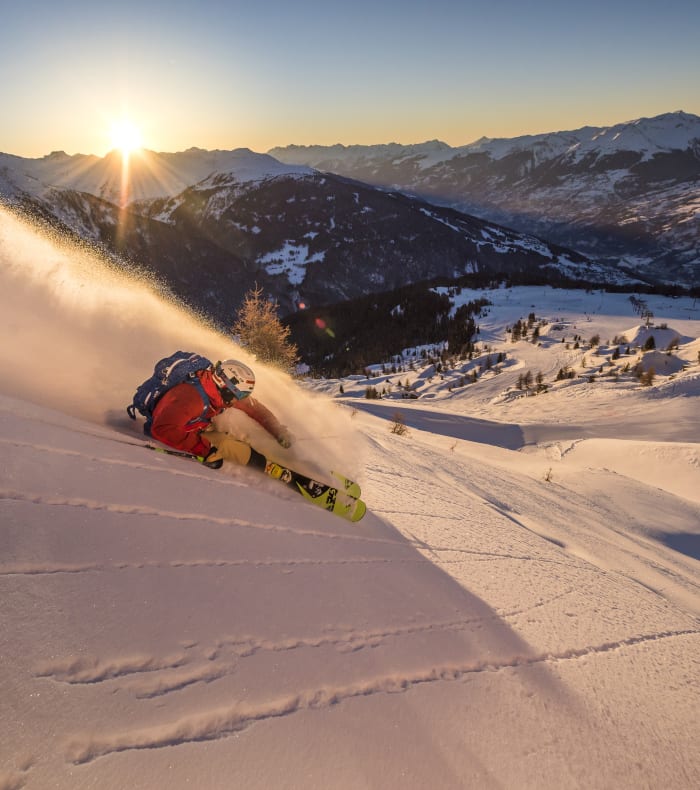 Skieur en rouge dévalant une pente hors-piste dans les Alpes avec vue panoramique sur la vallée enneigée au lever du soleil