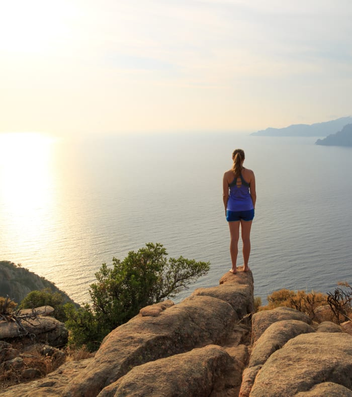 Randonneuse sur rochers face à la mer de Corse au coucher de soleil avec vue sur les montagnes