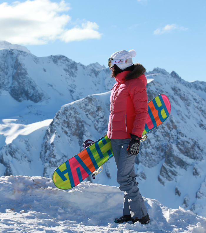 Snowboarder en veste rouge tenant sa planche colorée face aux montagnes enneigées et ciel bleu