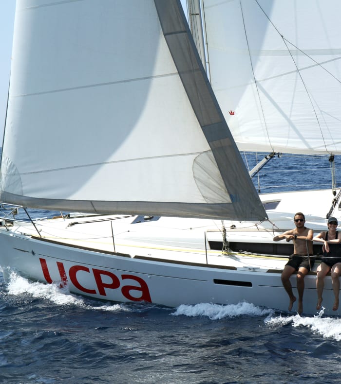 Voilier UCPA avec équipage naviguant en Morbihan, voiles blanches déployées sur mer bleue et côtes bretonnes en arrière-plan