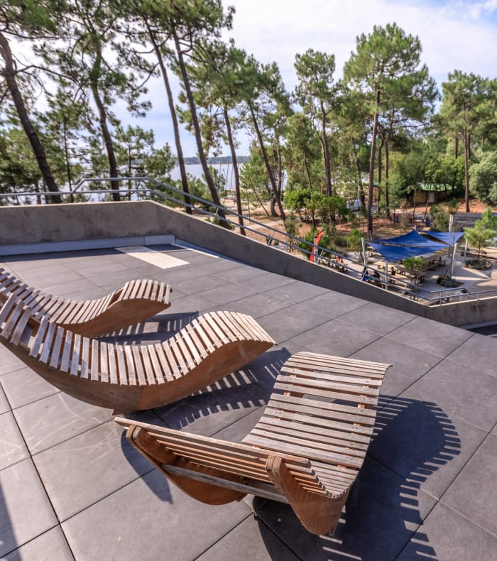 Terrasse avec chaises longues en bois face aux terrains de sport et lac de Bombannes entouré de pins