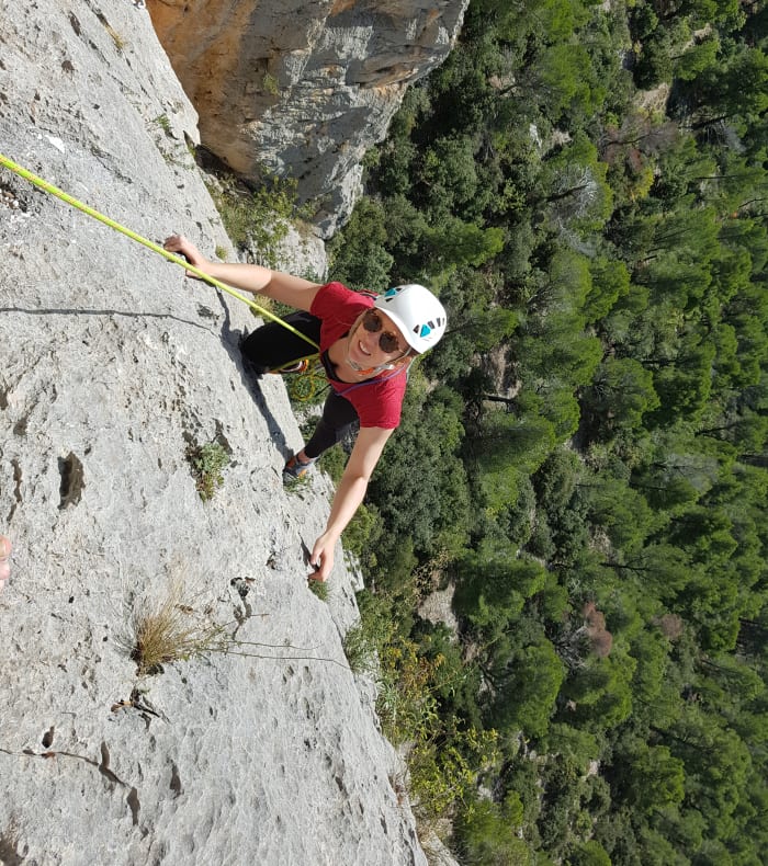 Participant en escalade sur paroi rocheuse avec casque et équipement de sécurité, forêt verdoyante en arrière-plan