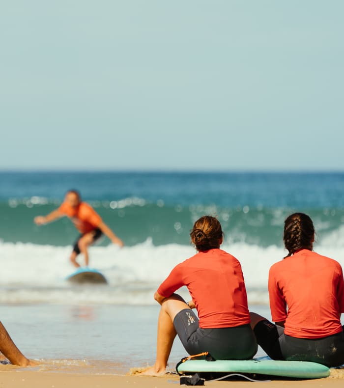 Groupe de surfeurs en combinaison orange à La Torche, certains dans l'océan et d'autres assis sur la plage avec leurs planches de surf